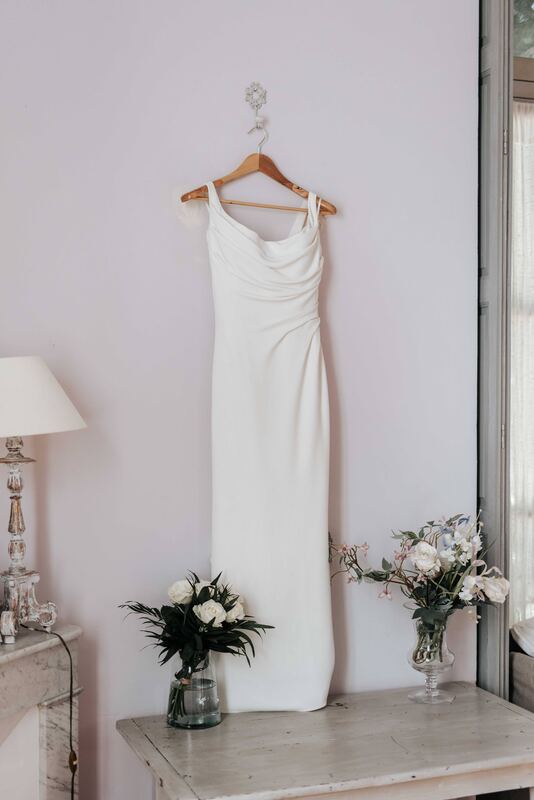 Robe de mariée suspendue dans une pièce claire, bouquets de fleurs.