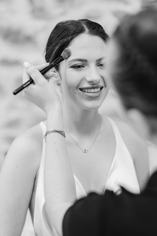 Une mariée rit pendant son maquillage, en noir et blanc.