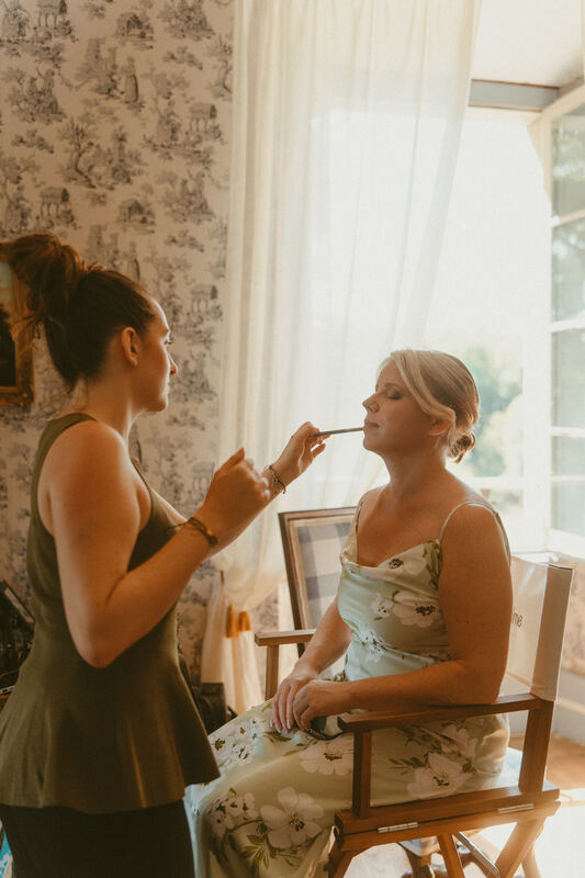 Margaux maquille une mariée assise près de la fenêtre.