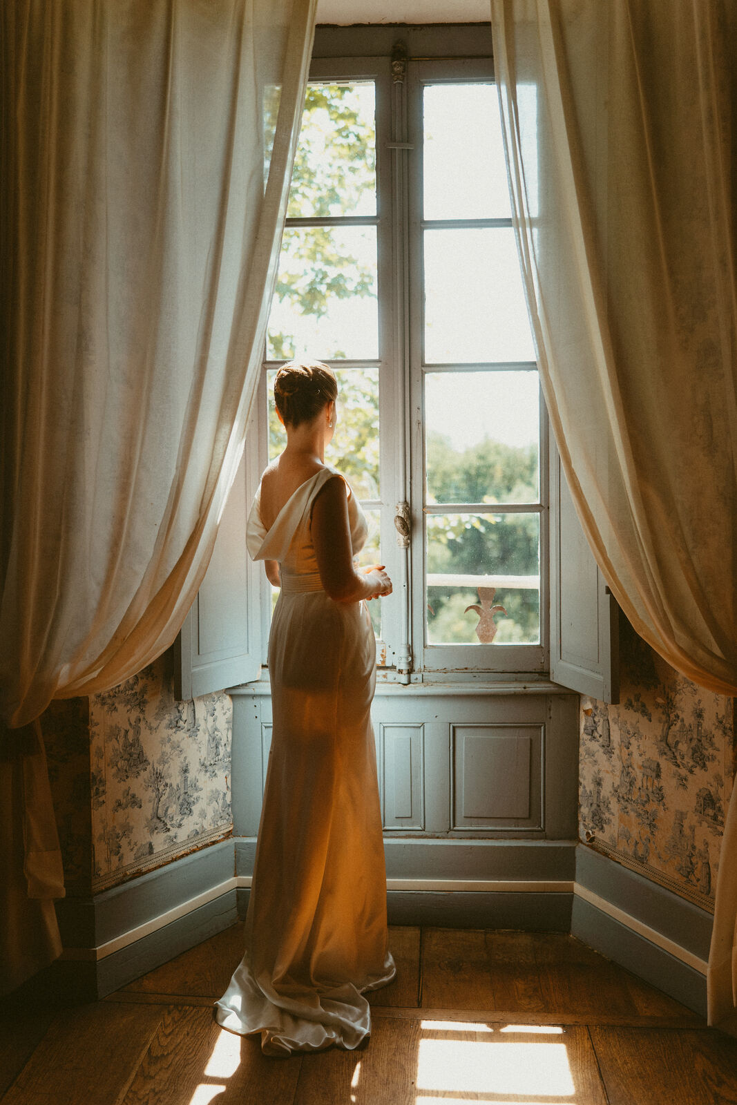Une mariée à la fenêtre, dans la lumière du matin, entre les rideaux.